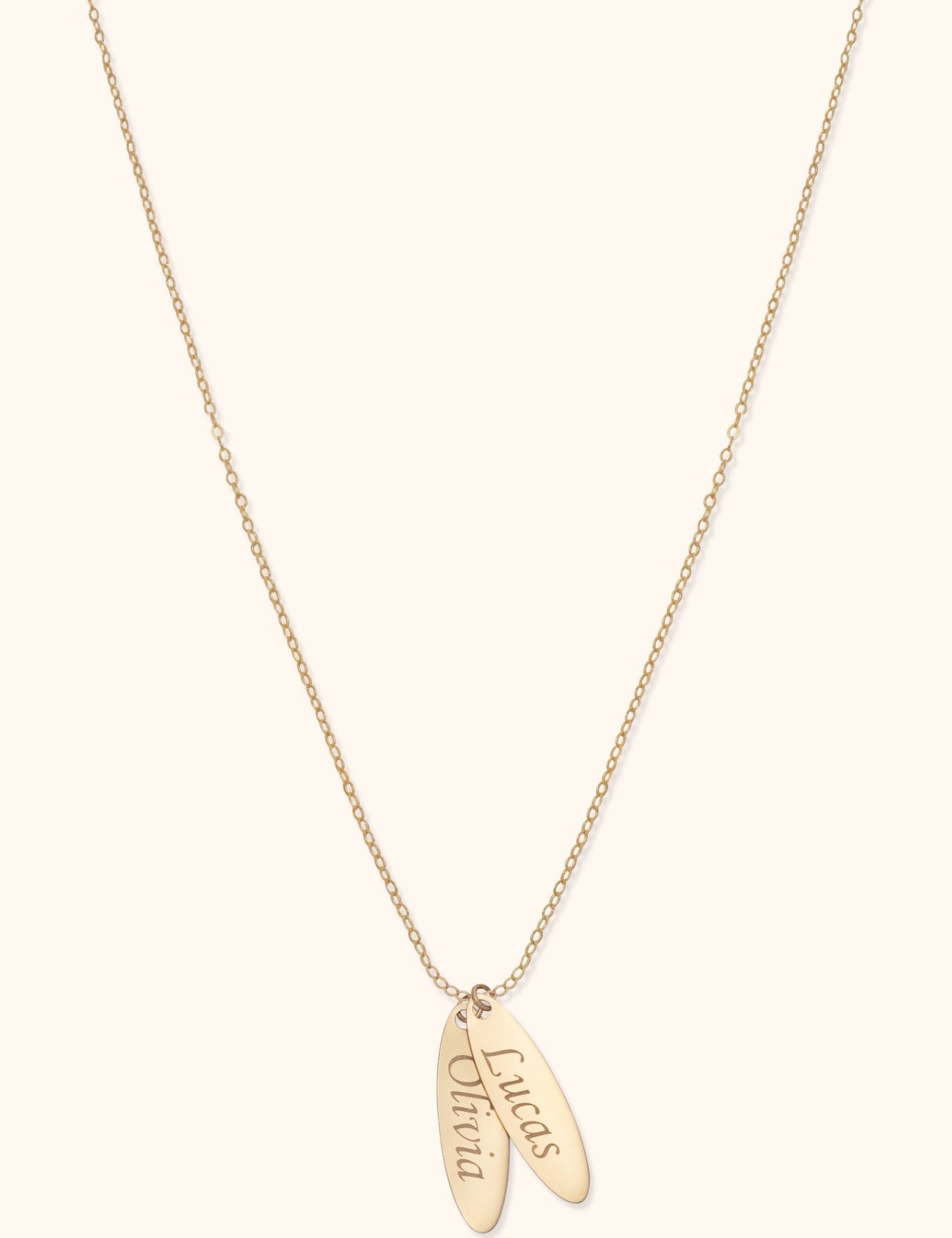 Gold-colored fine chain necklace with two overlapping elongated oval tags bearing the names Lucas and Olivia on a cream background.