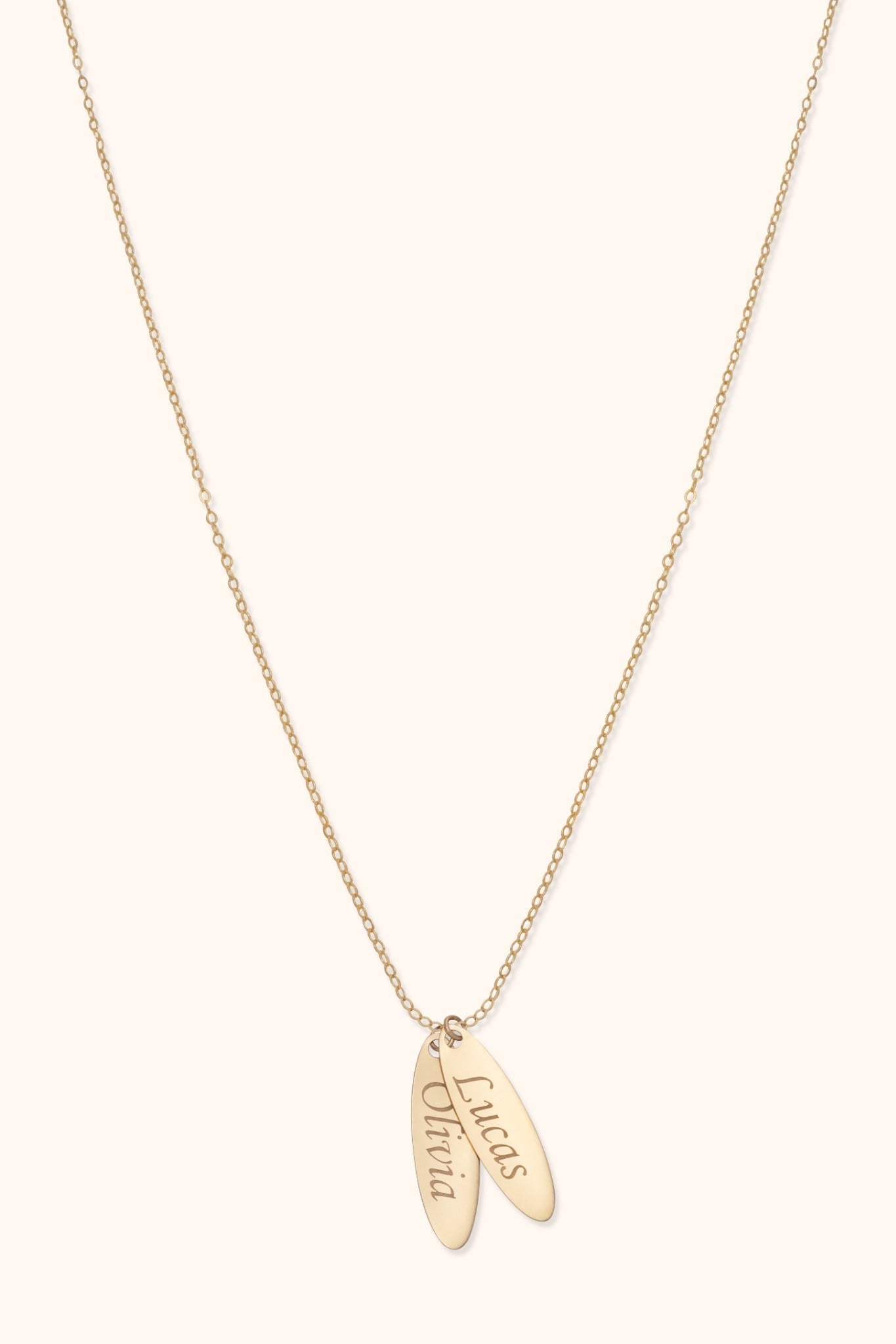 Gold-colored fine chain necklace with two overlapping elongated oval tags bearing the names Lucas and Olivia on a cream background.