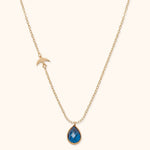Gold-toned chain necklace with a small bird detail on one side and a faceted blue teardrop pendant on a cream background.