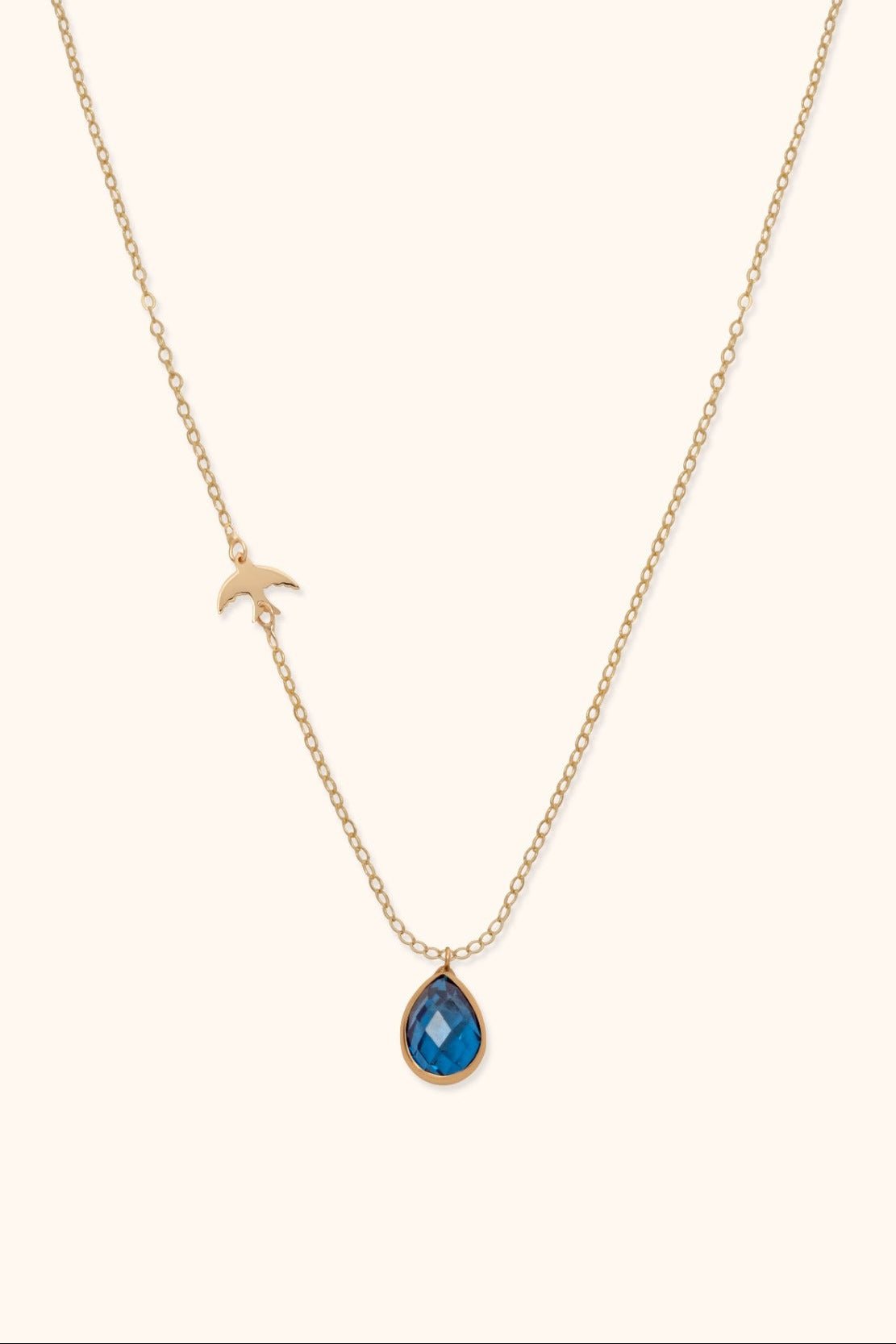 Gold-toned chain necklace with a small bird detail on one side and a faceted blue teardrop pendant on a cream background.