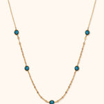 Gold-toned chain necklace with five round, faceted blue accents and small flattened links against a cream background.