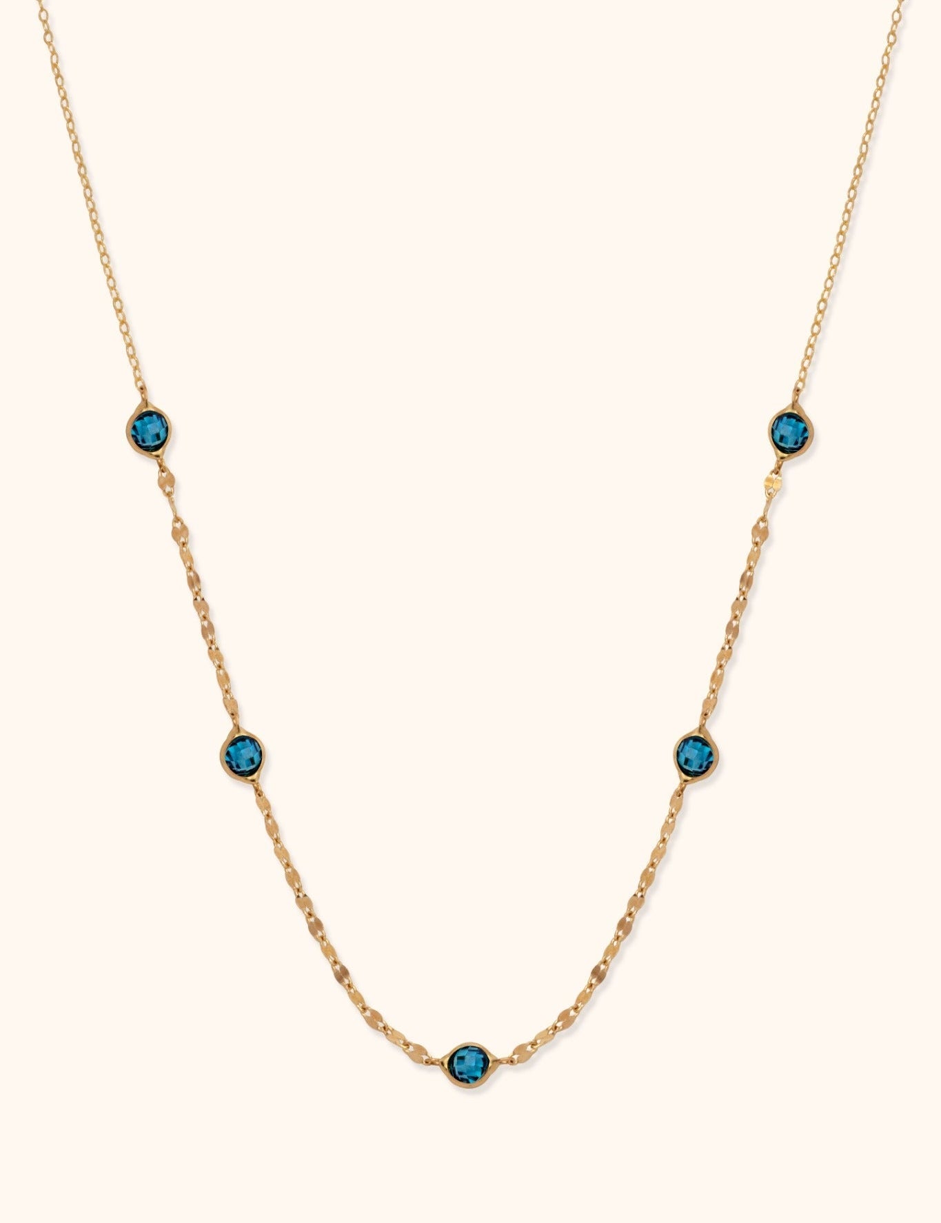Gold-toned chain necklace with five round, faceted blue accents and small flattened links against a cream background.
