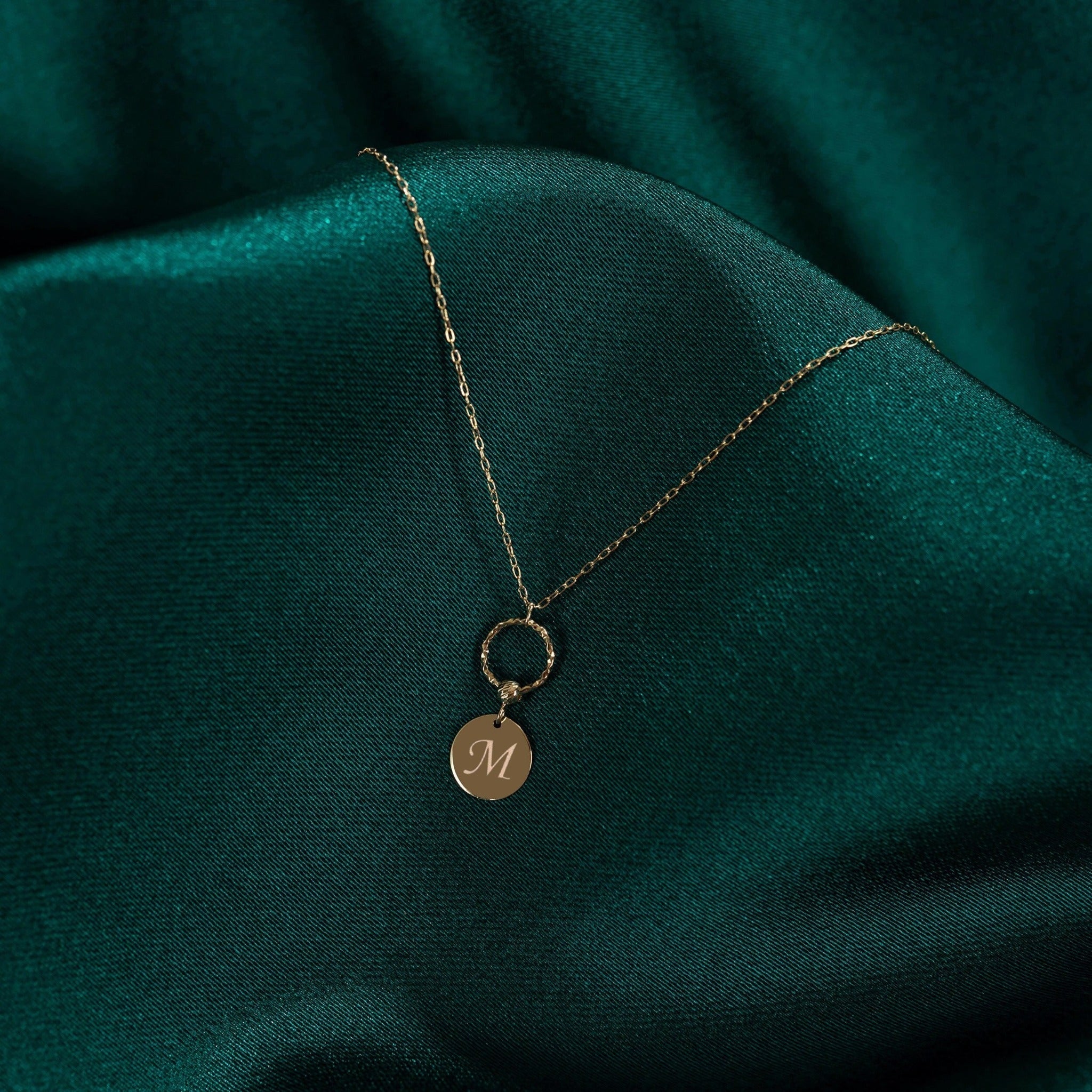 Gold-tone fine chain necklace with a textured ring and round disc bearing a script M, resting on dark green fabric.