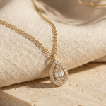 Gold-toned chain necklace with a teardrop pendant featuring clear round and rectangular accents, draped over cream fabric.