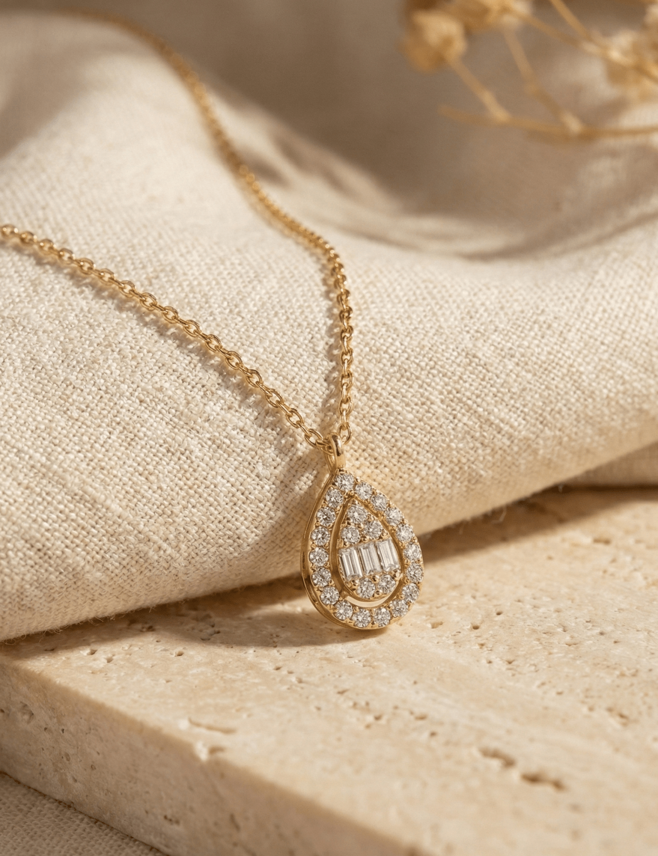 Gold-toned chain necklace with a teardrop pendant featuring clear round and rectangular accents, draped over cream fabric.