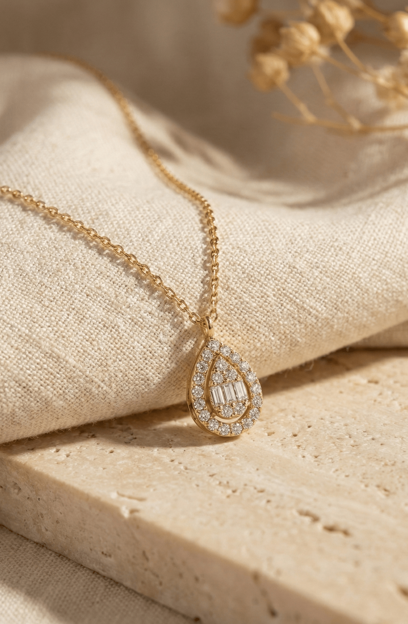 Gold-toned chain necklace with a teardrop pendant featuring clear round and rectangular accents, draped over cream fabric.