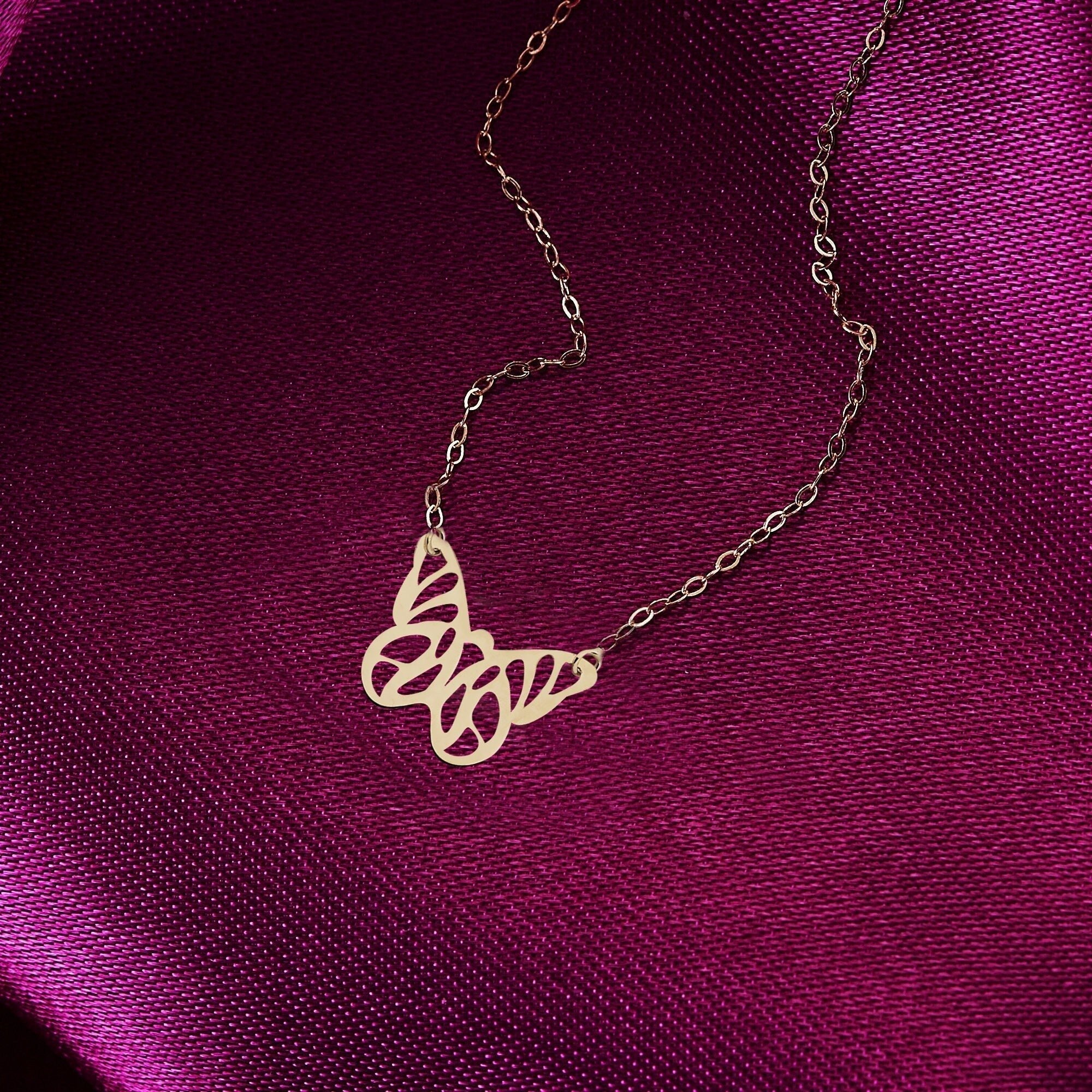 Gold-colored necklace with an openwork butterfly pendant and fine oval-link chain resting on deep magenta fabric.