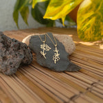 Two gold-toned necklaces with openwork flower pendants and delicate chains draped over a dark stone, with leafy plants behind.