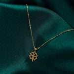 Gold-colored necklace with a four-leaf clover pendant featuring heart-shaped cutouts, resting on dark teal fabric.