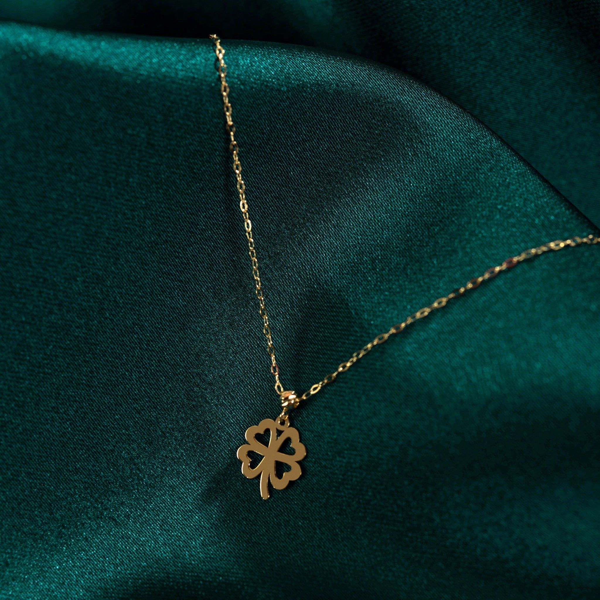 Gold-colored necklace with a four-leaf clover pendant featuring heart-shaped cutouts, resting on dark teal fabric.