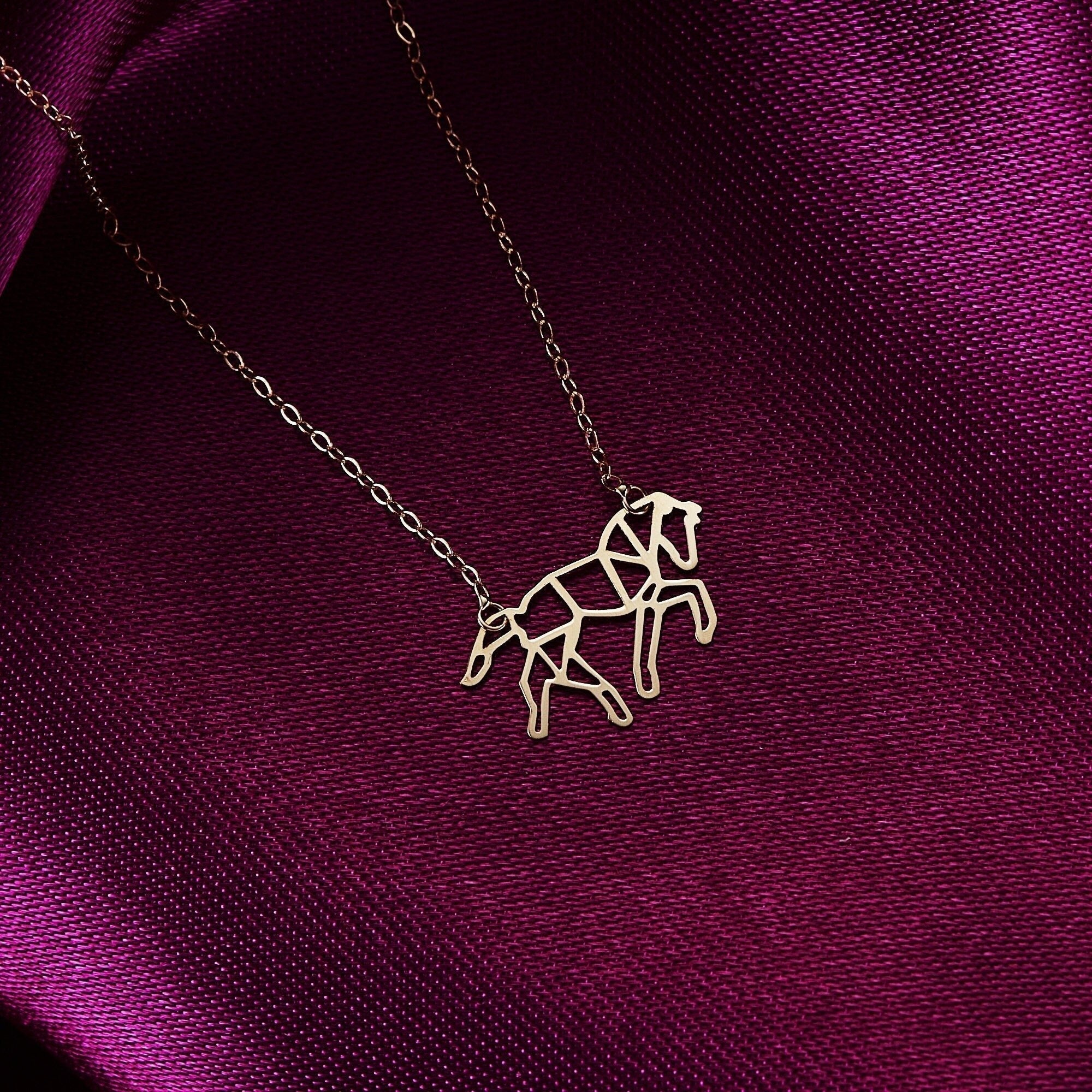 Gold-toned necklace with an openwork geometric horse pendant on a fine oval-link chain, resting on shiny purple fabric.