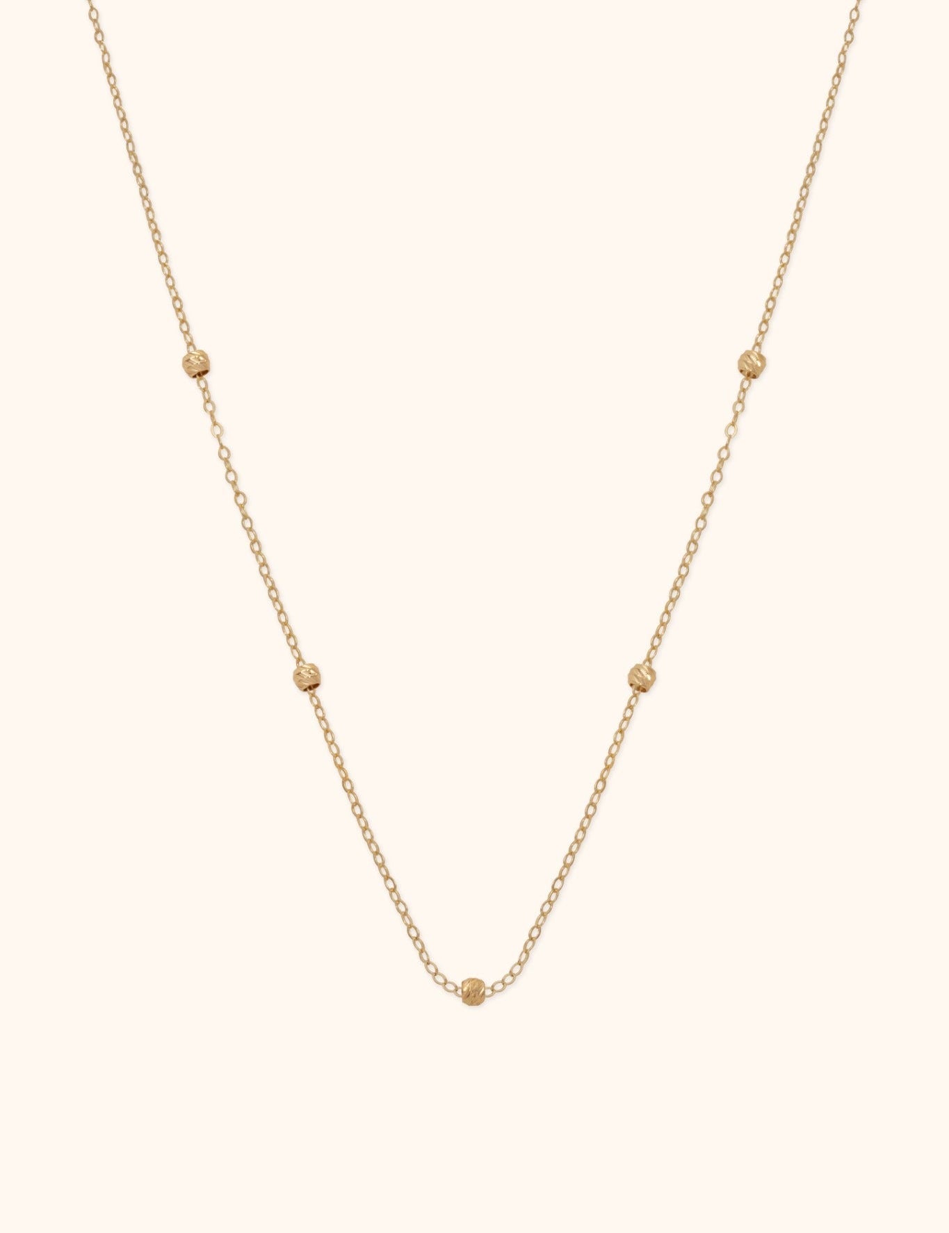 Gold-toned oval-link necklace with five small textured beads spaced along the chain, shown against a cream background.