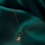 Gold-tone necklace with a fine oval-link chain, textured ring and flat tilted heart pendant on dark teal fabric.