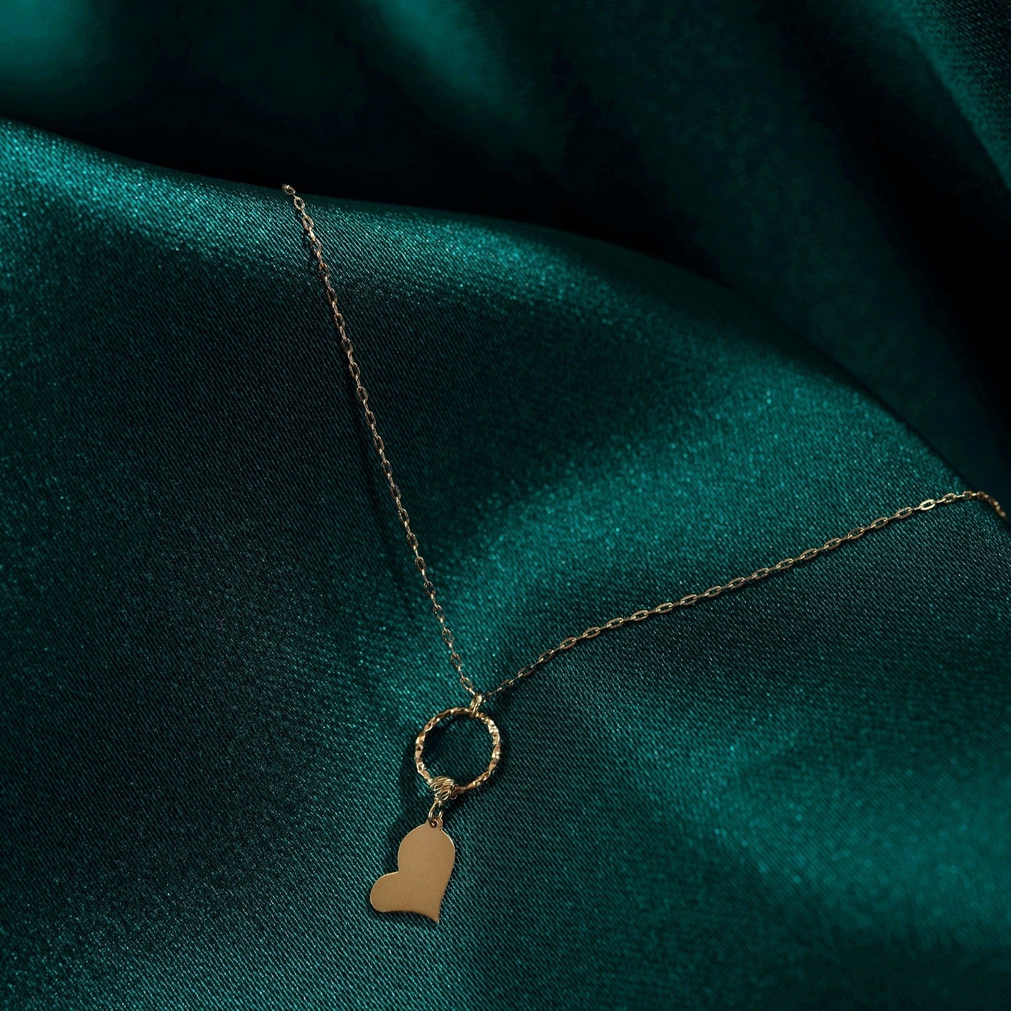 Gold-tone necklace with a fine oval-link chain, textured ring and flat tilted heart pendant on dark teal fabric.