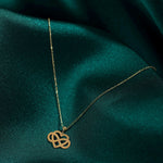 Gold-toned necklace with a fine oval-link chain and an intertwined heart and infinity pendant on dark teal fabric.