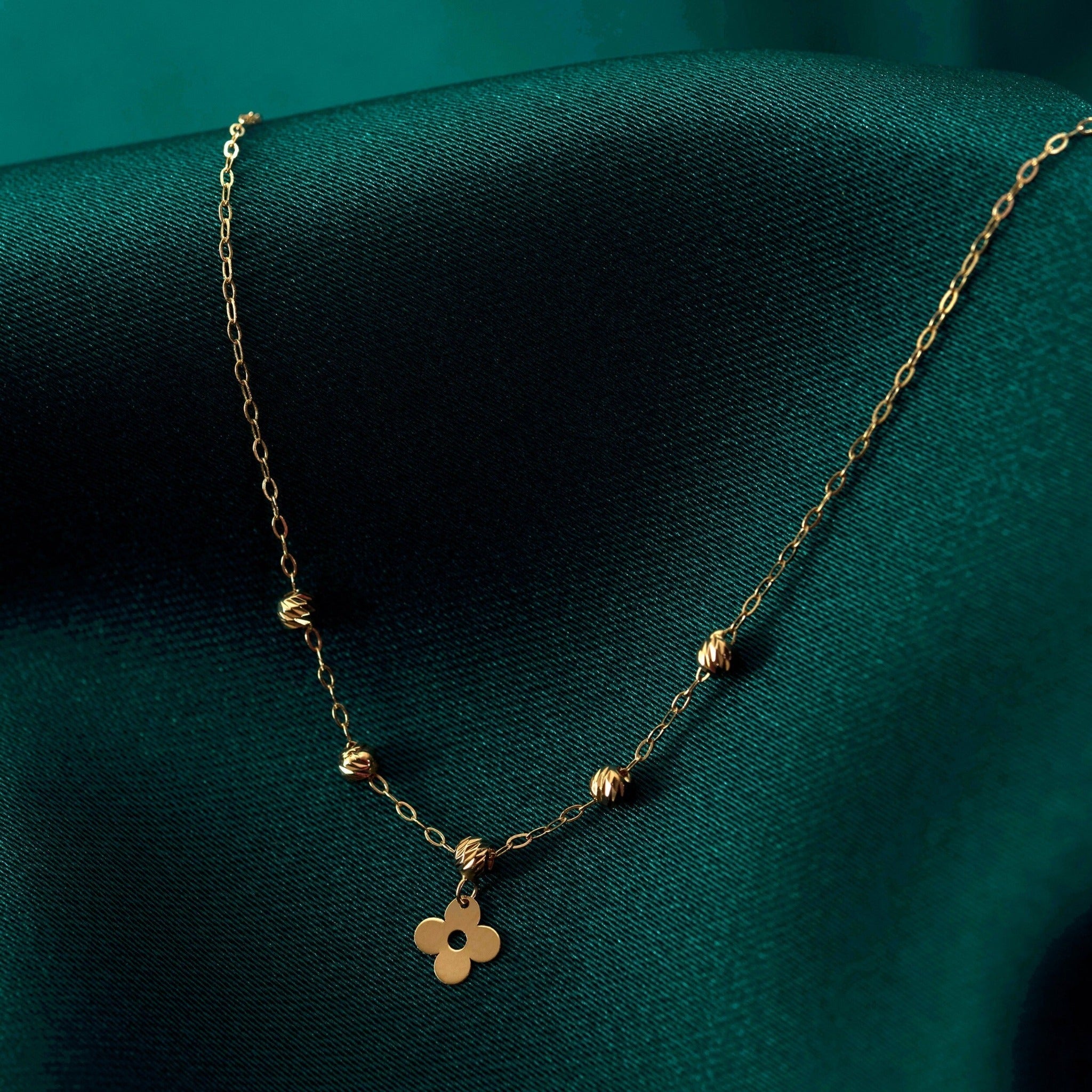 Gold-colored oval-link necklace with five textured beads and a four-lobed charm with a central hole, resting on dark teal fabric.