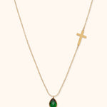 Gold-toned chain necklace with a cross on one side and a faceted green teardrop pendant on a cream background.