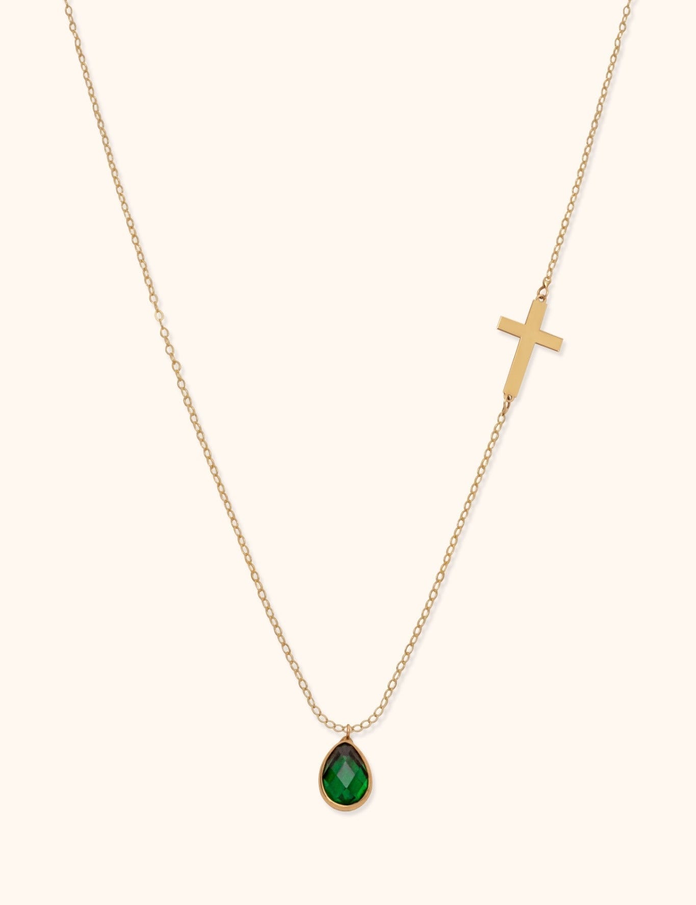 Gold-toned chain necklace with a cross on one side and a faceted green teardrop pendant on a cream background.