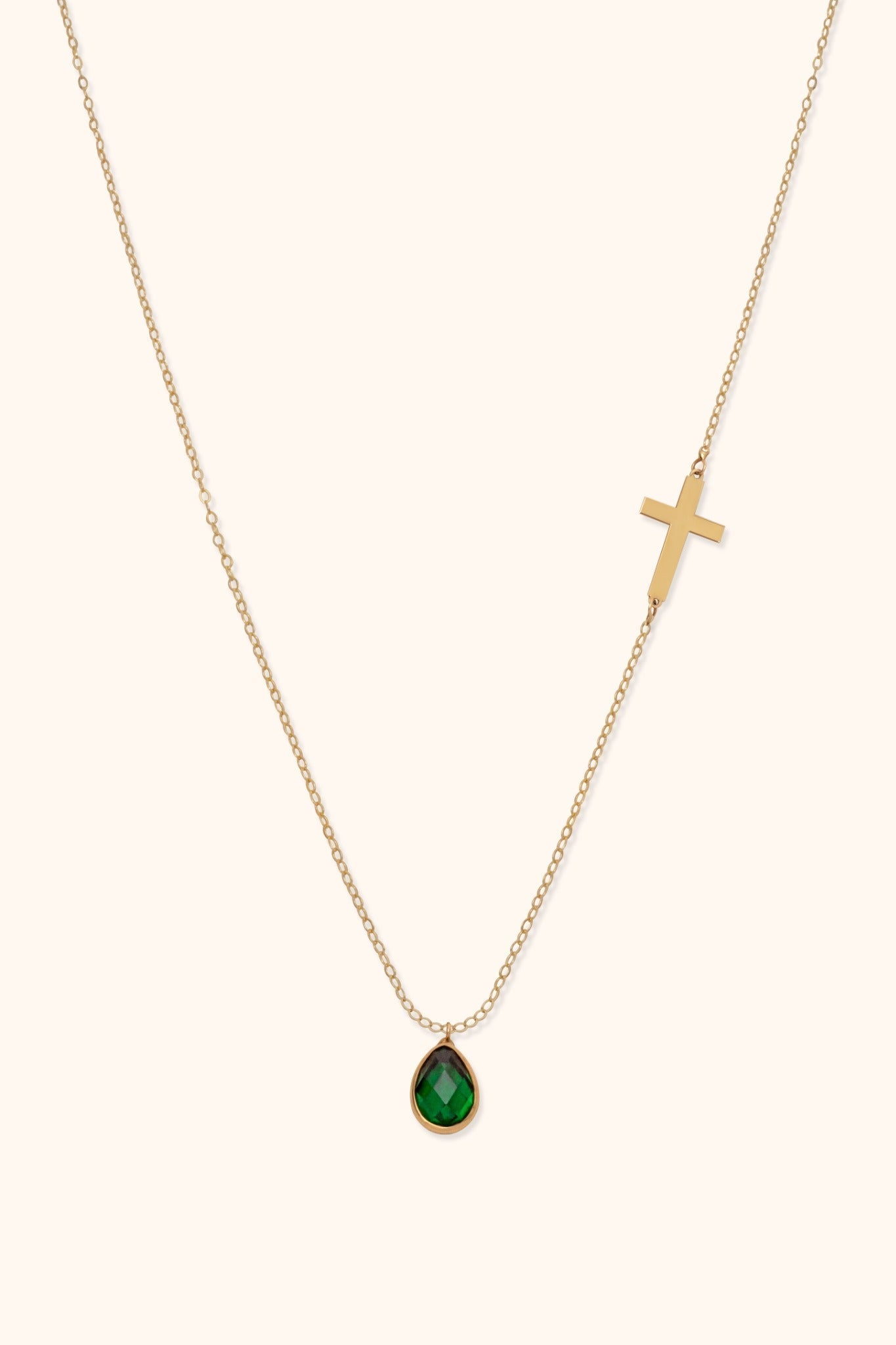 Gold-toned chain necklace with a cross on one side and a faceted green teardrop pendant on a cream background.