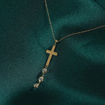 Gold-tone chain necklace with a flat cross pendant and three clear faceted dangles on dark teal fabric.