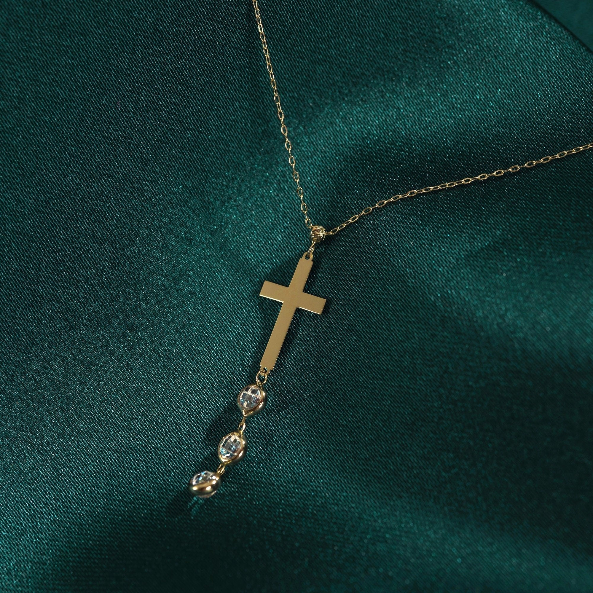 Gold-tone chain necklace with a flat cross pendant and three clear faceted dangles on dark teal fabric.