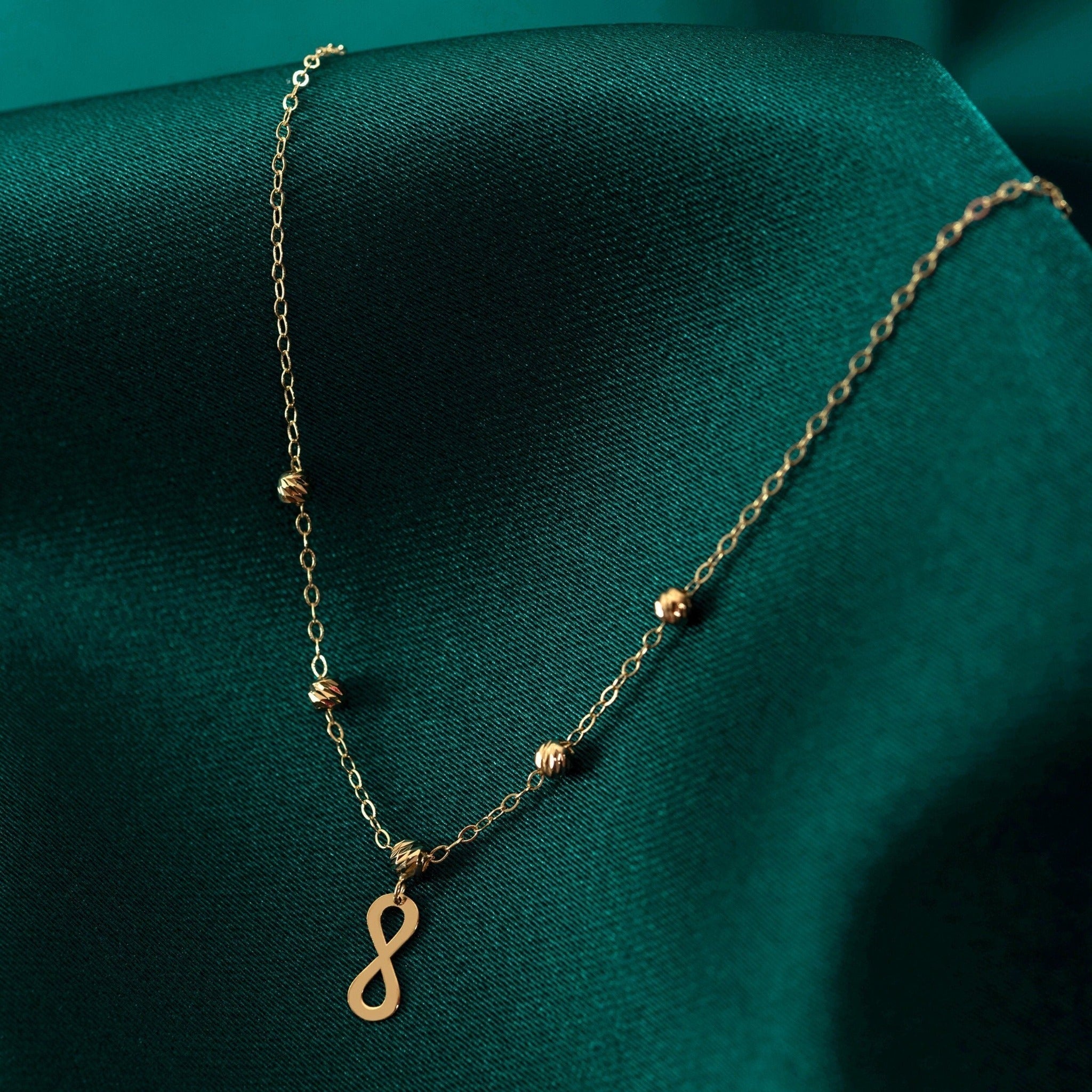 Gold-toned necklace with an infinity pendant and textured round beads along a fine oval-link chain on dark teal fabric.