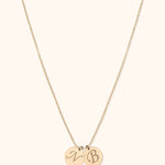 Gold-toned fine chain necklace with two overlapping round pendants bearing decorative initials on a cream background.