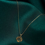 Gold-colored necklace with a fine oval-link chain and an openwork seashell pendant resting on dark teal fabric.