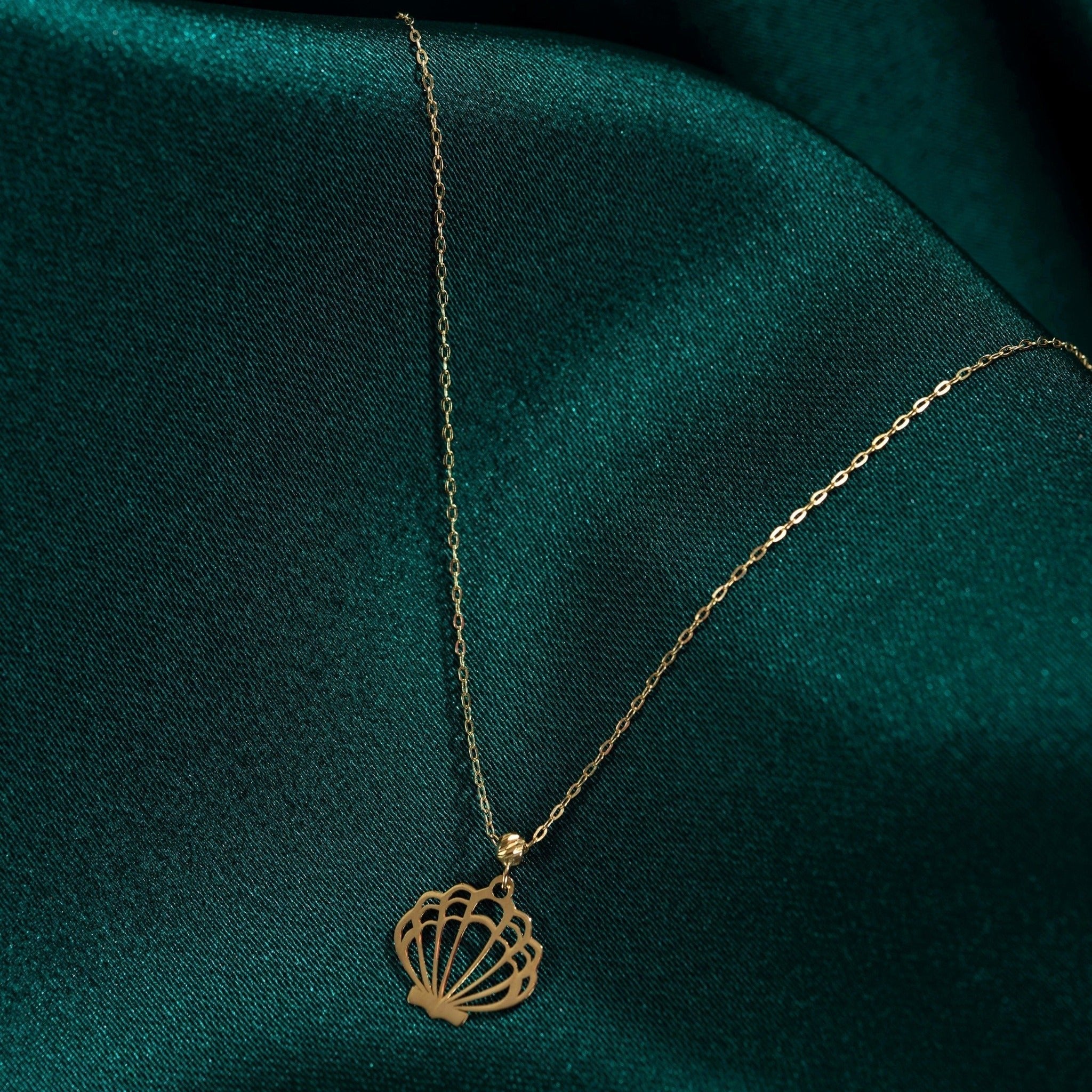 Gold-colored necklace with a fine oval-link chain and an openwork seashell pendant resting on dark teal fabric.