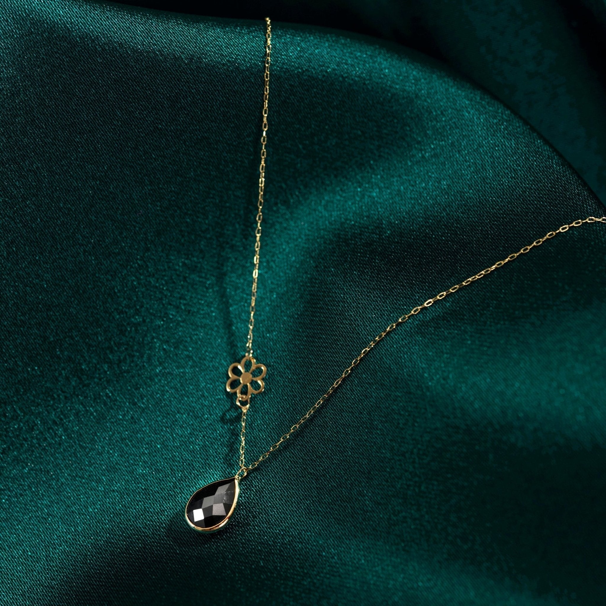 Gold-tone chain necklace with an open flower charm and faceted black teardrop pendant on dark green fabric.