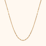 Thin gold-tone necklace with small, reflective oval links, arranged in a long U shape against a cream background.