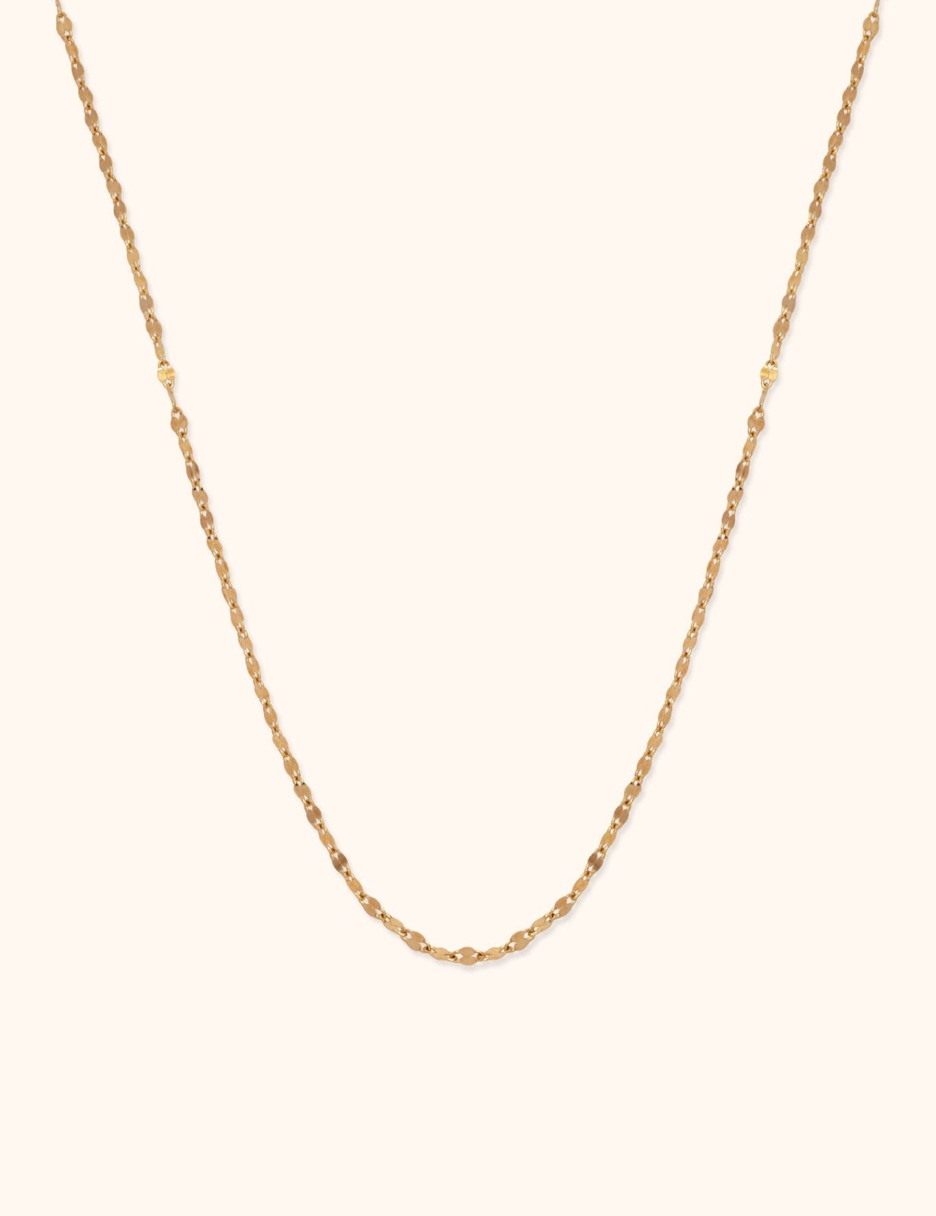 Thin gold-tone necklace with small, reflective oval links, arranged in a long U shape against a cream background.