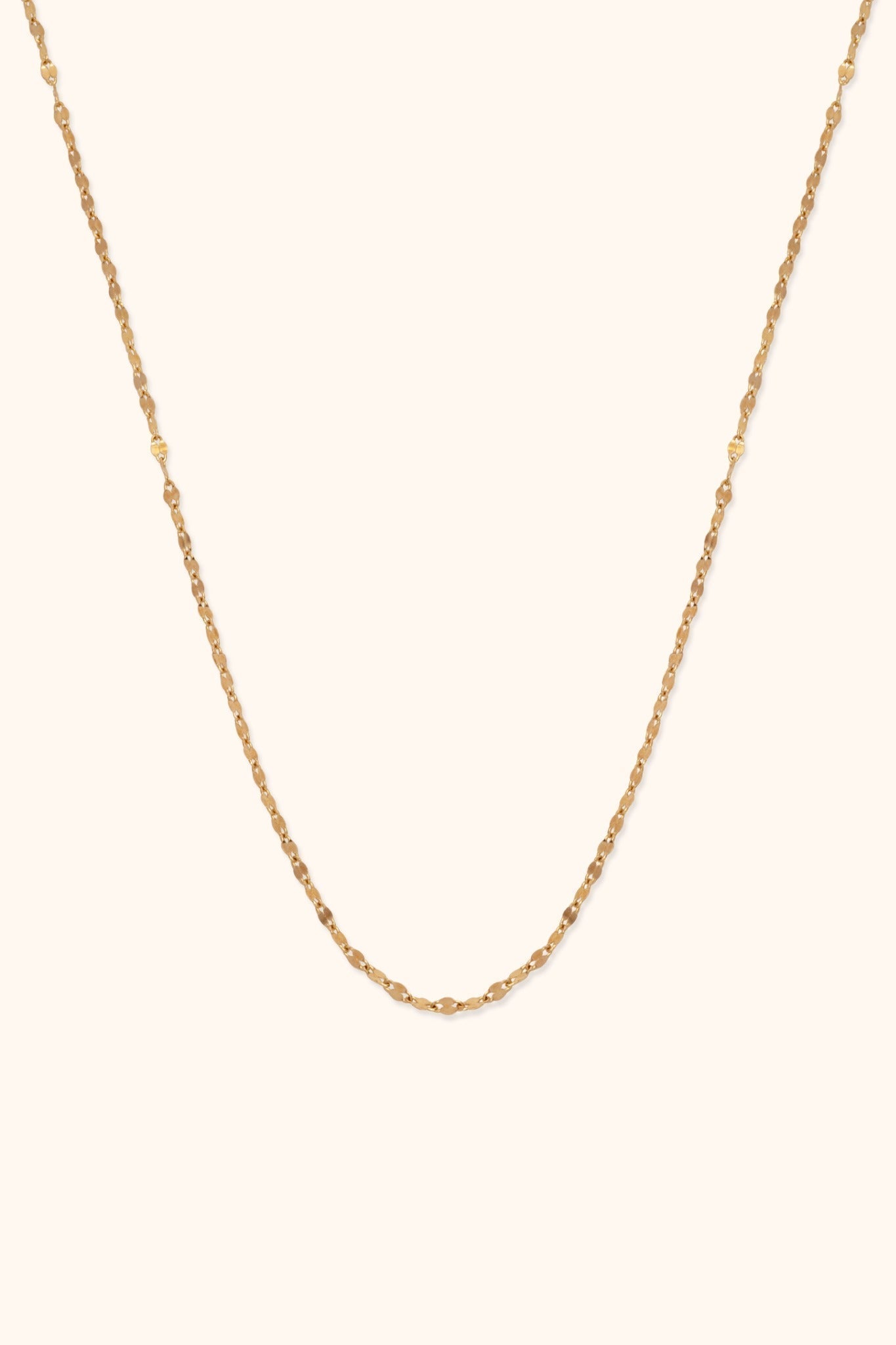 Thin gold-tone necklace with small, reflective oval links, arranged in a long U shape against a cream background.