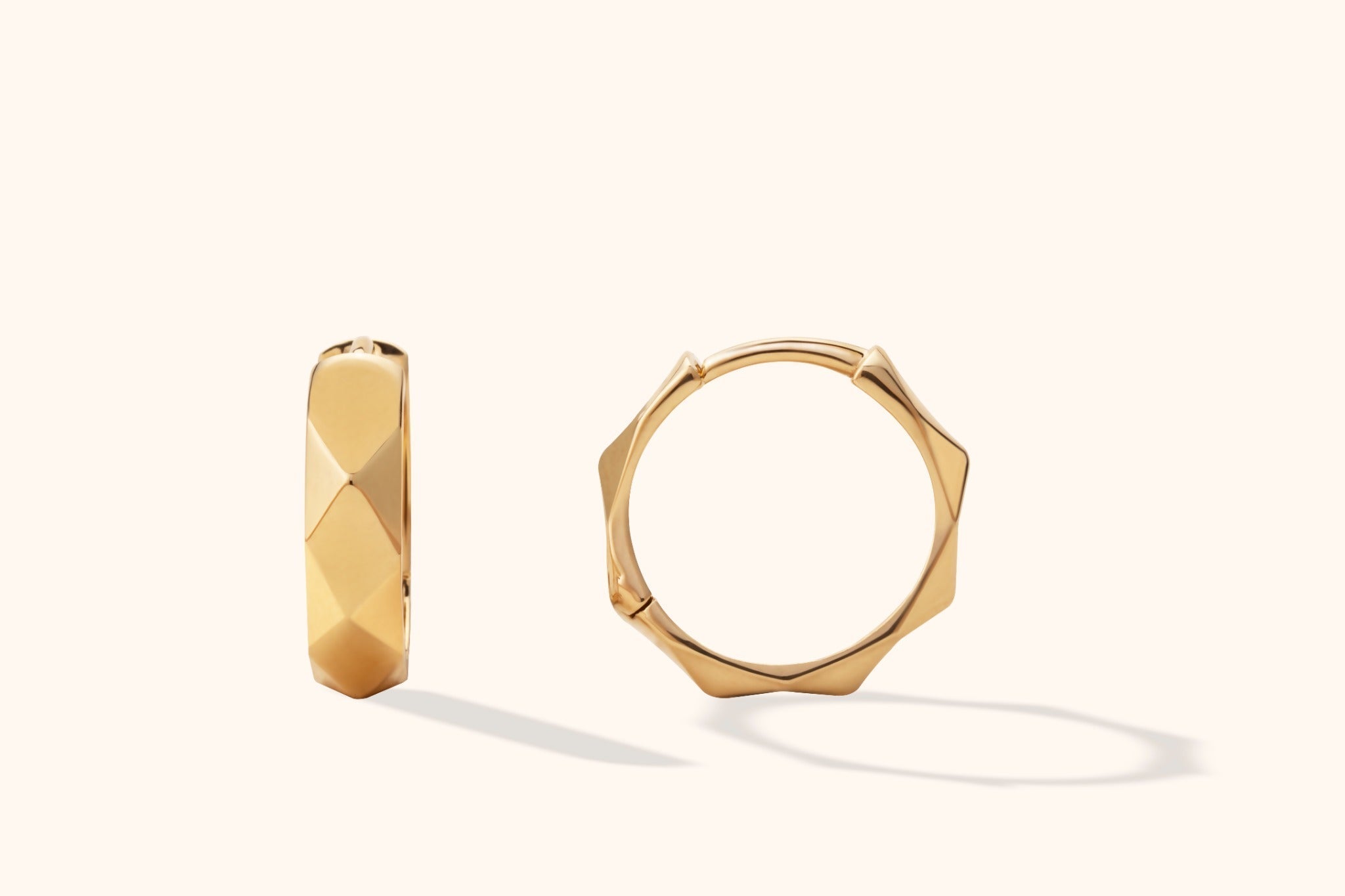 Two gold-colored hoop earrings with angular, faceted surfaces, shown from different angles against a cream background.