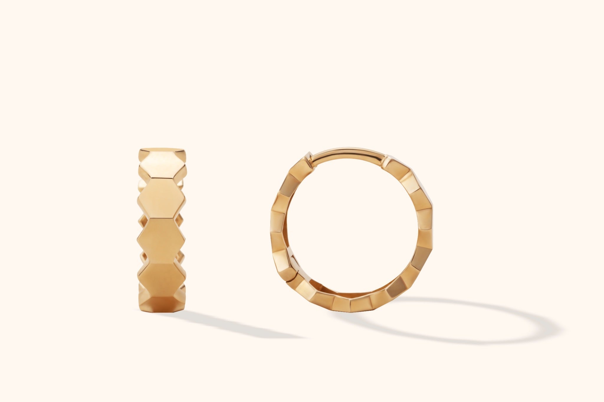 Two gold-toned hoop earrings with polished hexagonal facets, shown from different angles on a pale cream background.
