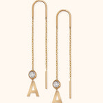 Pair of gold-colored threader earrings with long chains, clear faceted accents, and dangling letter A charms on a cream background.