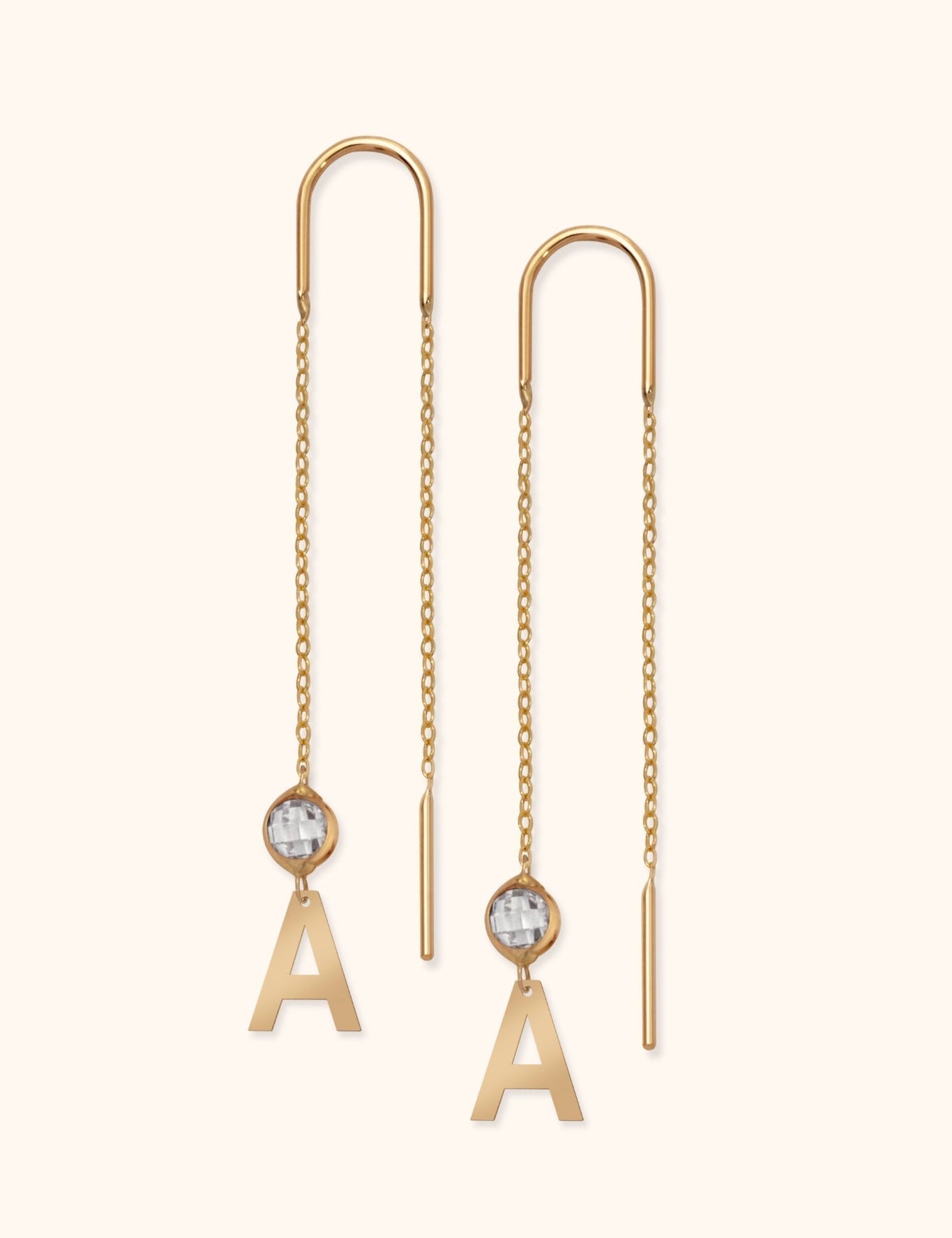 Pair of gold-colored threader earrings with long chains, clear faceted accents, and dangling letter A charms on a cream background.