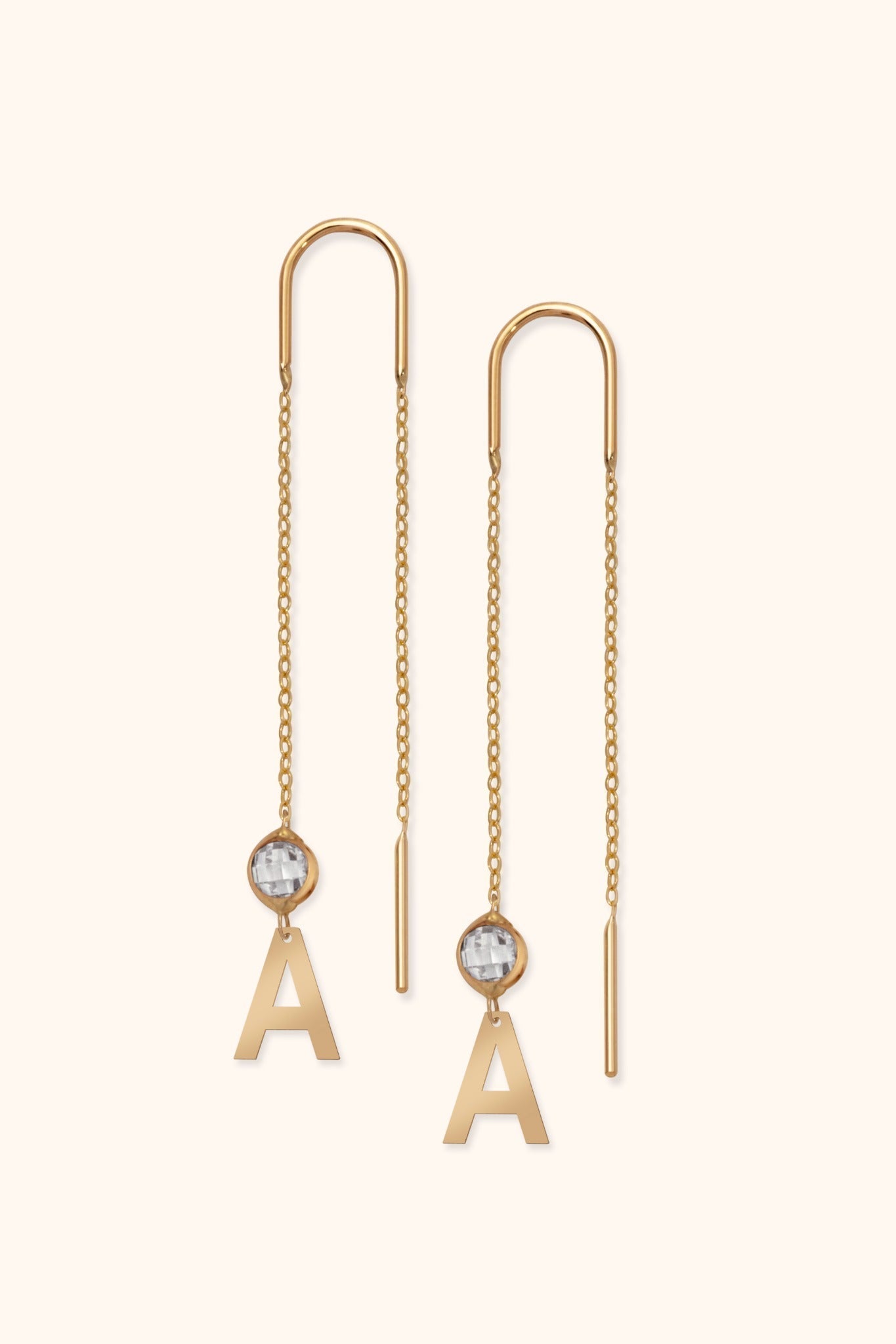 Pair of gold-colored threader earrings with long chains, clear faceted accents, and dangling letter A charms on a cream background.