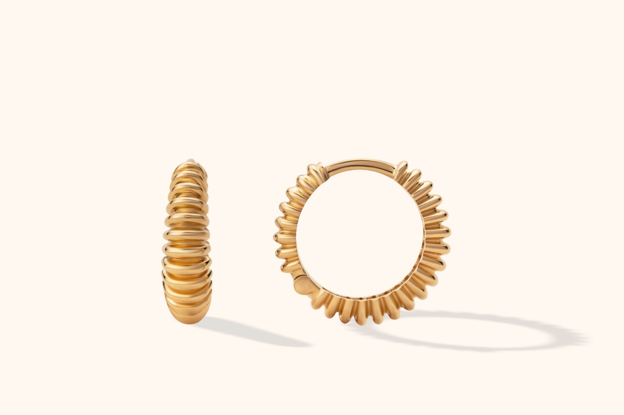 Two gold-colored hoop earrings with rounded ribs, shown from different angles on a pale cream background.
