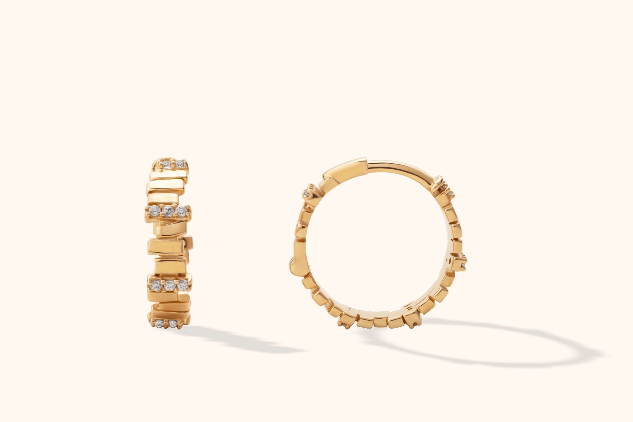 Two gold-tone hoop earrings with raised rectangular bars and rows of small clear stones, shown from different angles on a cream background.
