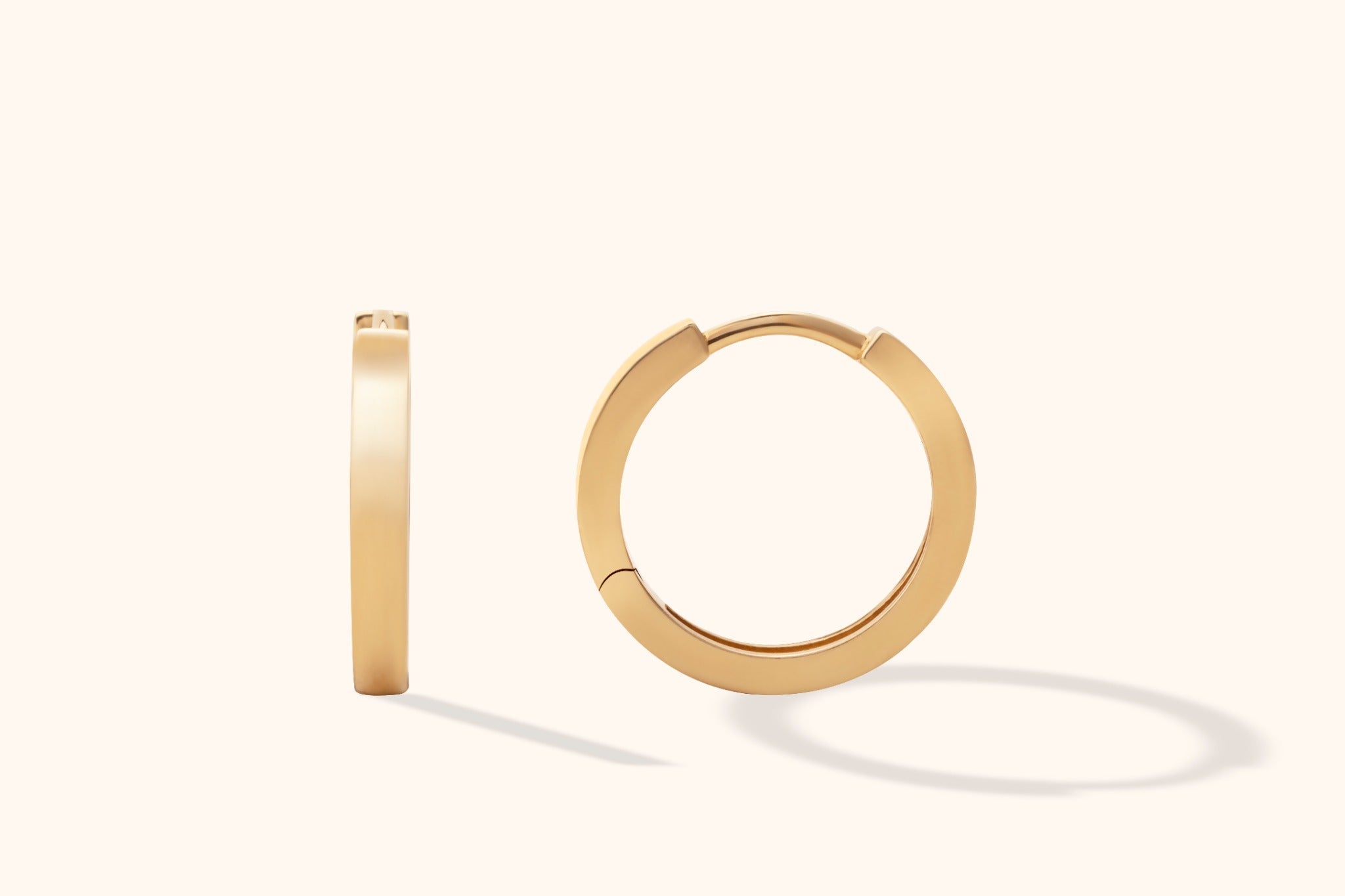 Two smooth gold-colored hoop earrings, one viewed edge-on and the other showing its circular shape, on a cream background.