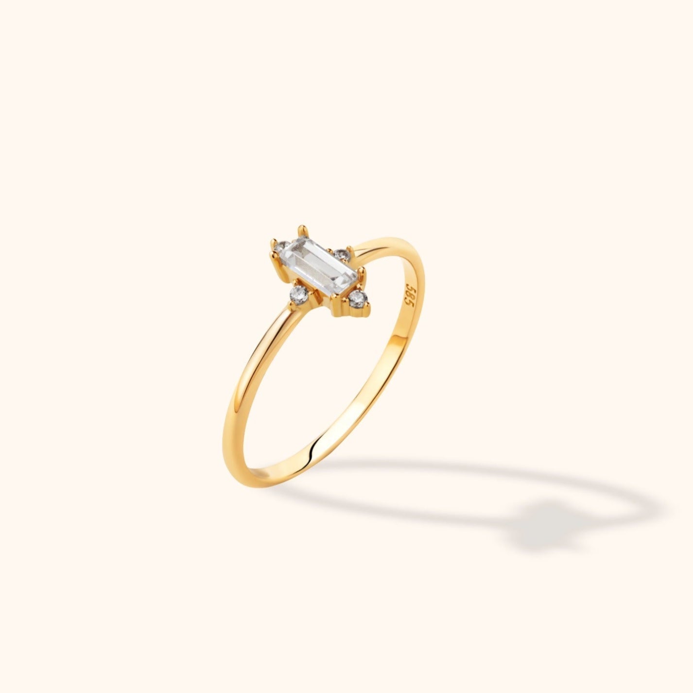 Gold-toned ring with a slim polished band, a clear rectangular center stone and small round stones, shown at an angle.