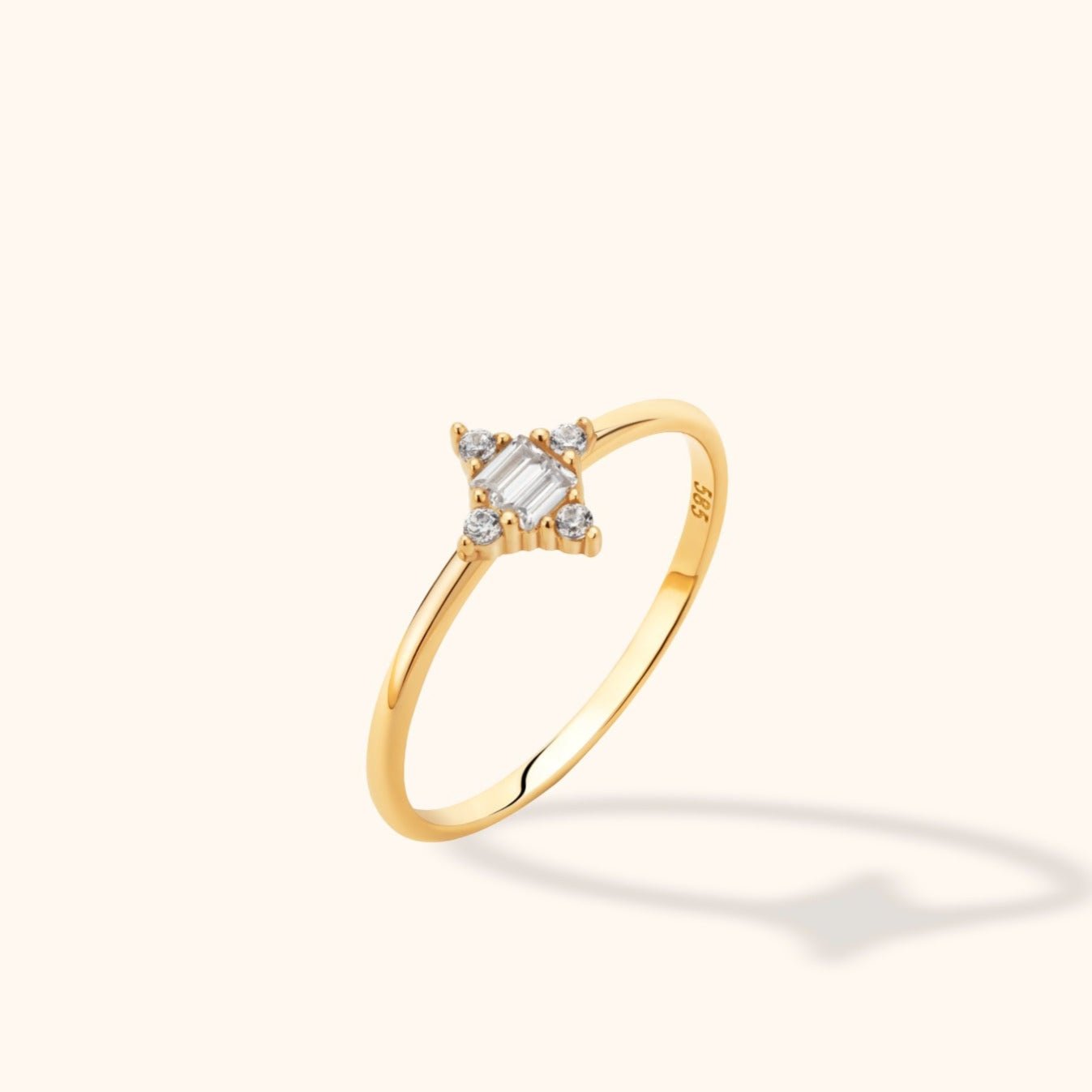 Slim gold-toned ring with a clear rectangular center stone and four round accents, shown at an angle on a cream background.