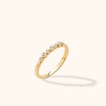 Slim gold-toned ring with alternating small and large round clear stones in bezel settings, tilted against a cream background.