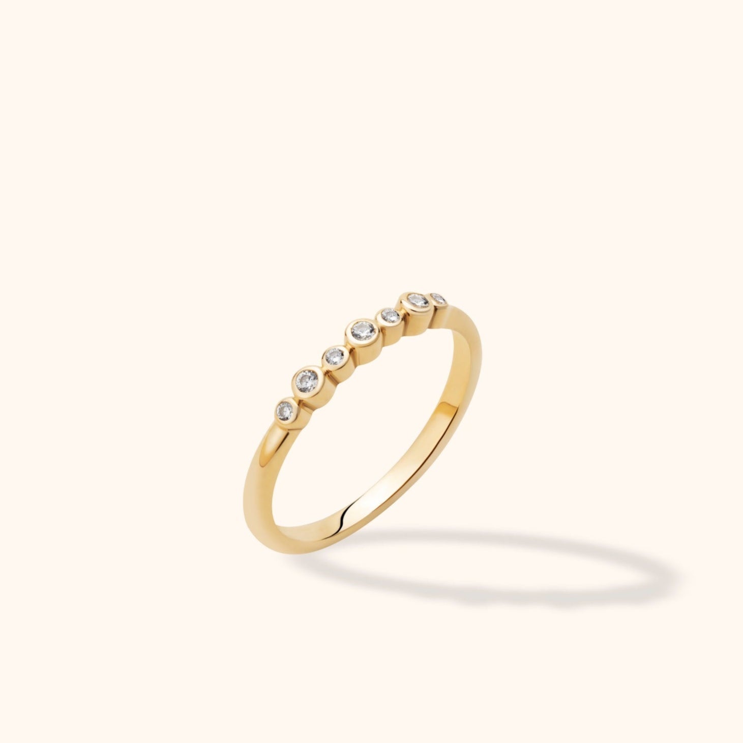 Slim gold-toned ring with alternating small and large round clear stones in bezel settings, tilted against a cream background.