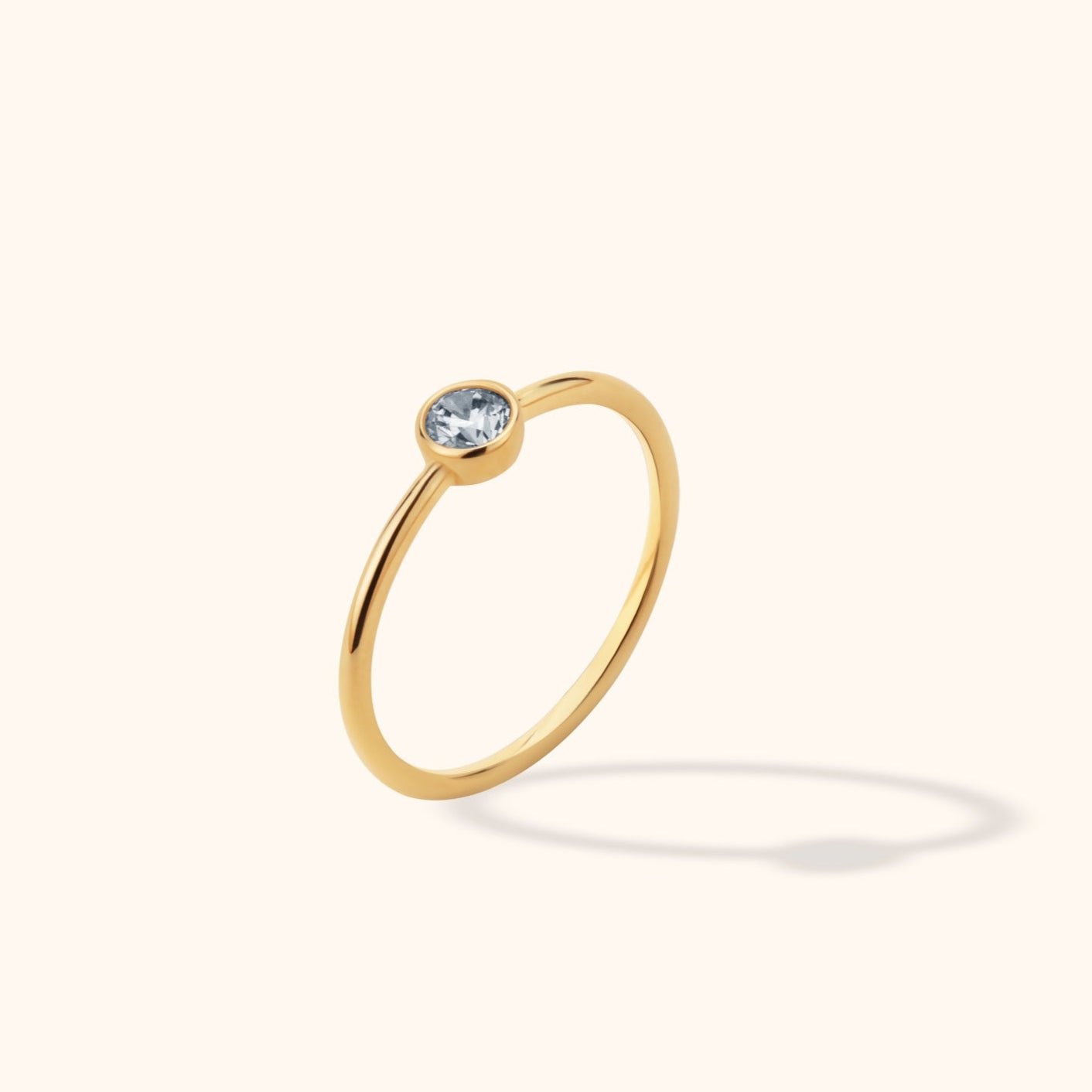 Slim gold-colored ring with a round, clear faceted stone in a bezel setting, shown tilted against a cream background.