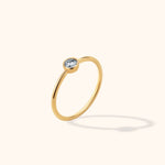 Slim gold-colored ring with a round, clear faceted stone in a bezel setting, shown tilted against a cream background.