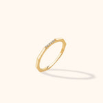 Gold-toned ring with angular edges and a short row of clear stones, tilted against a cream background with a soft shadow.