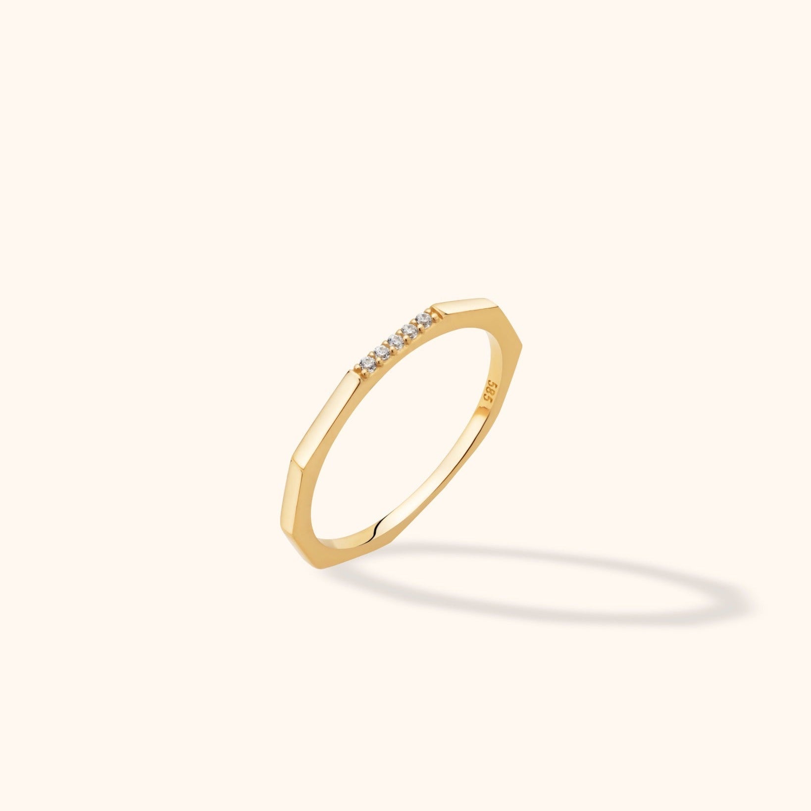 Gold-toned ring with angular edges and a short row of clear stones, tilted against a cream background with a soft shadow.