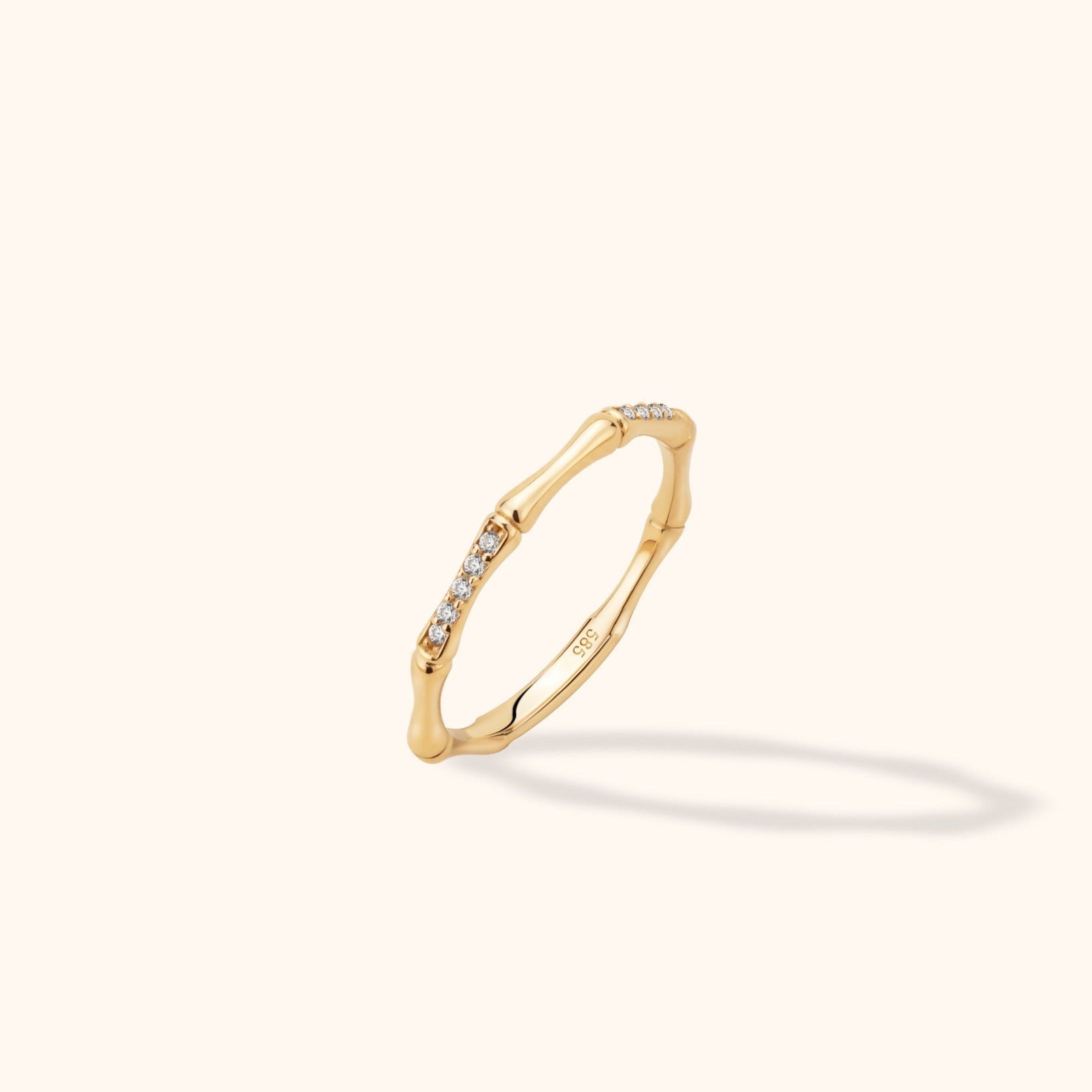 Gold-toned ring with a slender bamboo-like band and rows of small clear stones, shown tilted on a cream background.